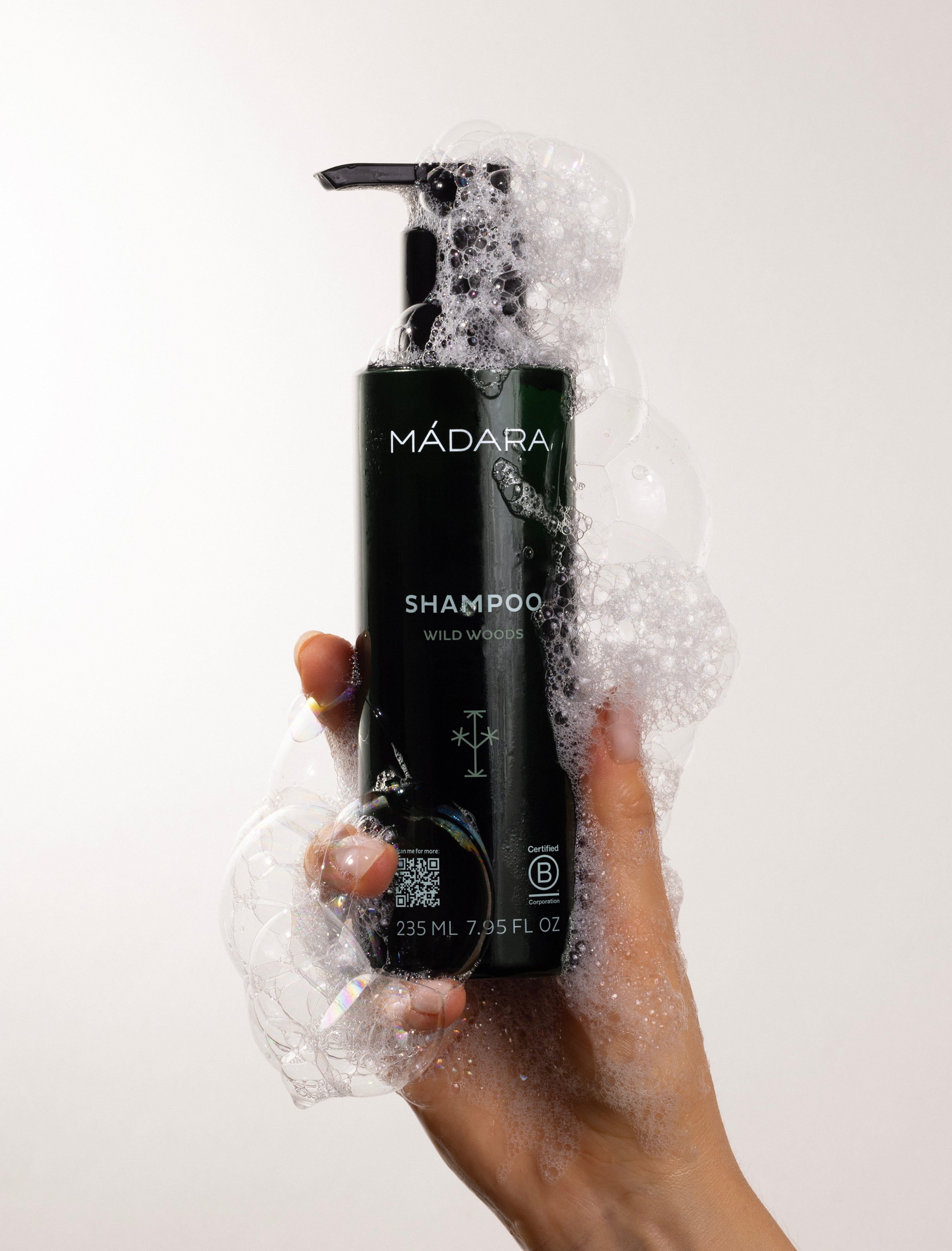 Mádara shampoo bottle with pump being held in a hand surrounded by foam on a white background